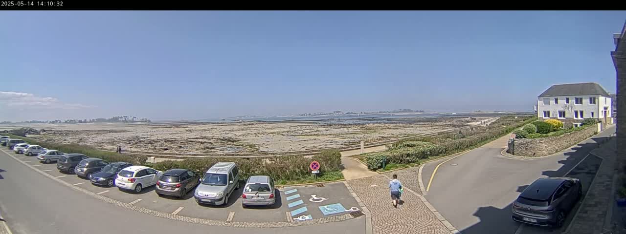 A sunny day reveals a coastal scene with several cars parked near a building, a person walking on a paved path, and a wide expanse of rocky shoreline extending to a distant landmass under a clear blue sky.