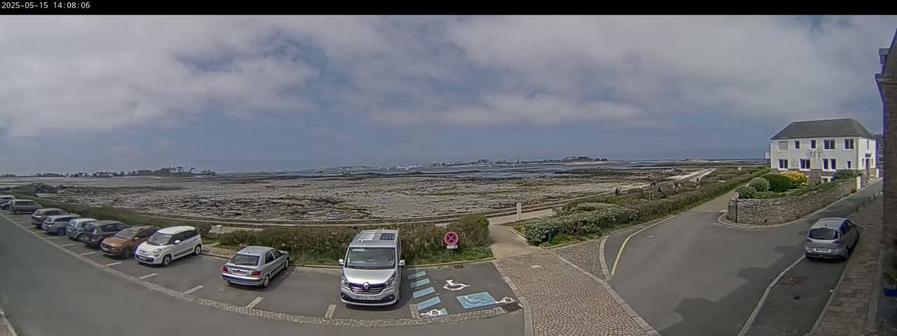 A mostly cloudy day reveals a coastal scene with a parking lot of cars near a stone building and a rocky shoreline extending to a distant landmass.