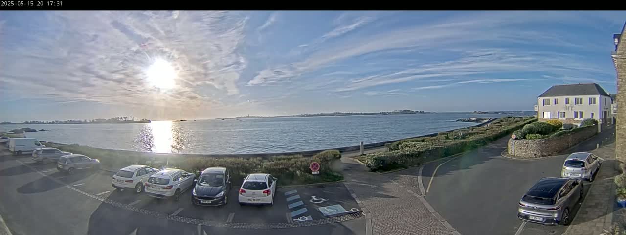 A partly sunny day shows a calm body of water with several cars parked near a building, with distant land visible across the water.