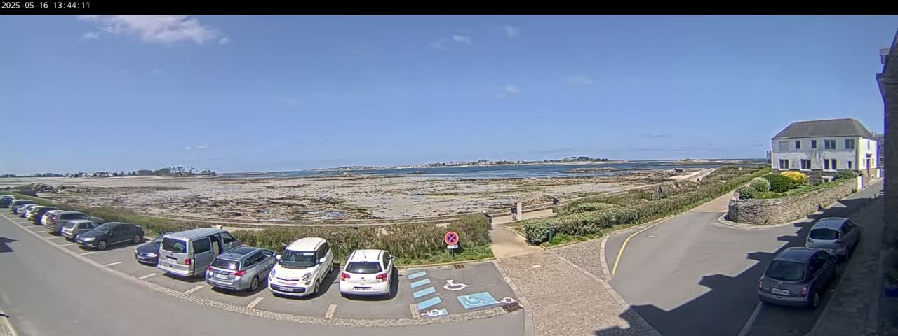Under a clear blue sky, several cars are parked in a lot overlooking a rocky shoreline and calm body of water, with a building visible in the upper right corner.