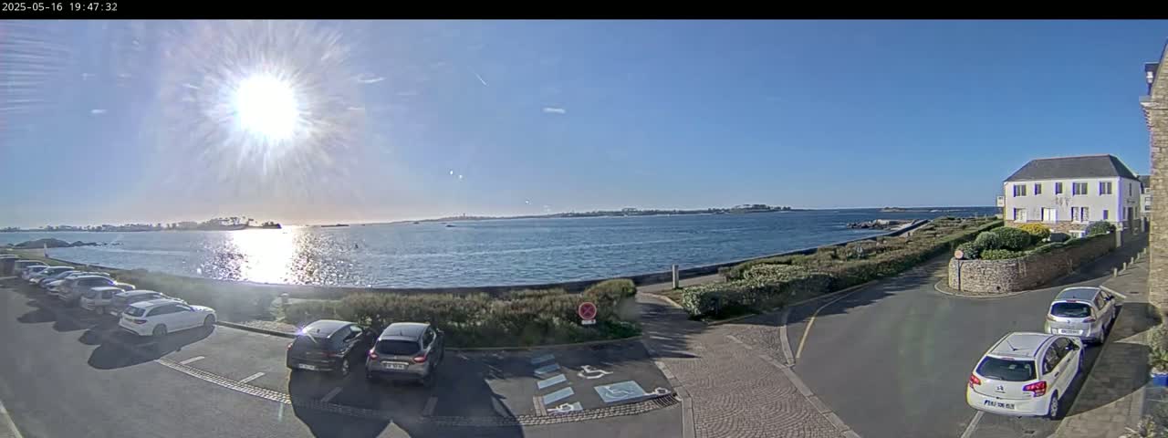 A sunny day overlooks a calm body of water, with parked cars in a lot next to a road and a building, all near a shoreline.