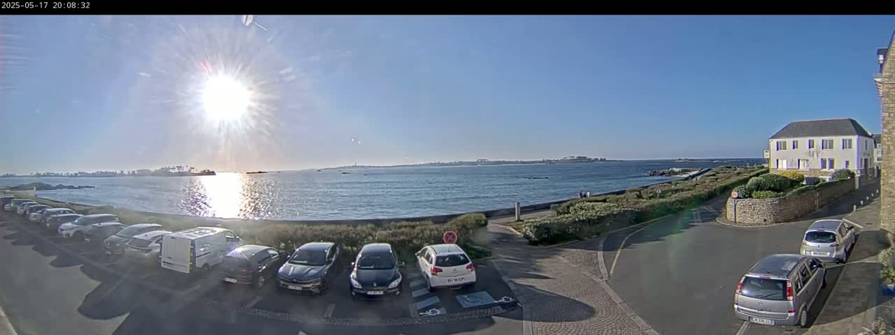 A sunny day reveals a seaside parking lot with several cars parked near a calm body of water, beyond which lies a distant coastline under a clear blue sky.