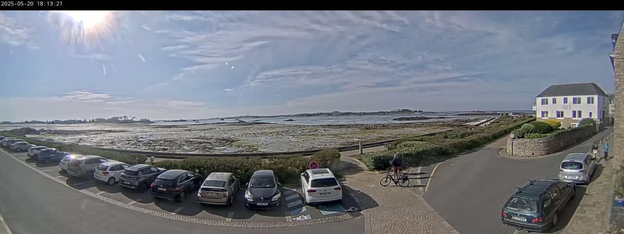 A sunny day reveals a coastal scene with a parking lot full of cars near a building, a calm body of water with exposed shorelines, and two people biking.
