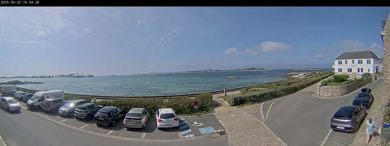 A sunny day overlooks a calm body of water with parked cars in a lot beside a building and a low stone wall.