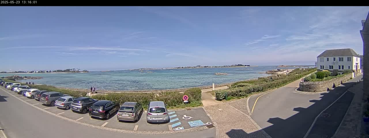 A sunny day reveals a calm body of water, a parking lot filled with cars, and a white building near a seaside path.