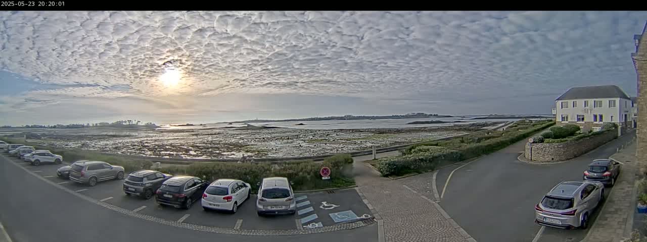 A mostly cloudy sunset over a calm body of water, with several cars parked in a lot next to a building and a low wall.
