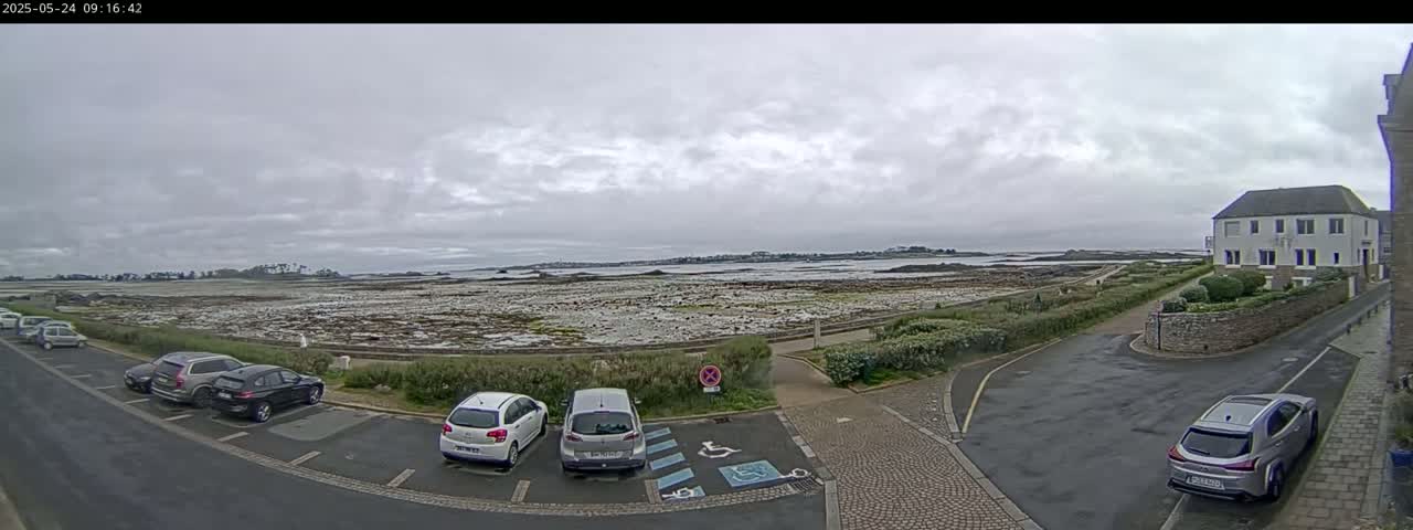 A parking lot with several cars overlooks a rocky, low-tide coastline under a cloudy sky.