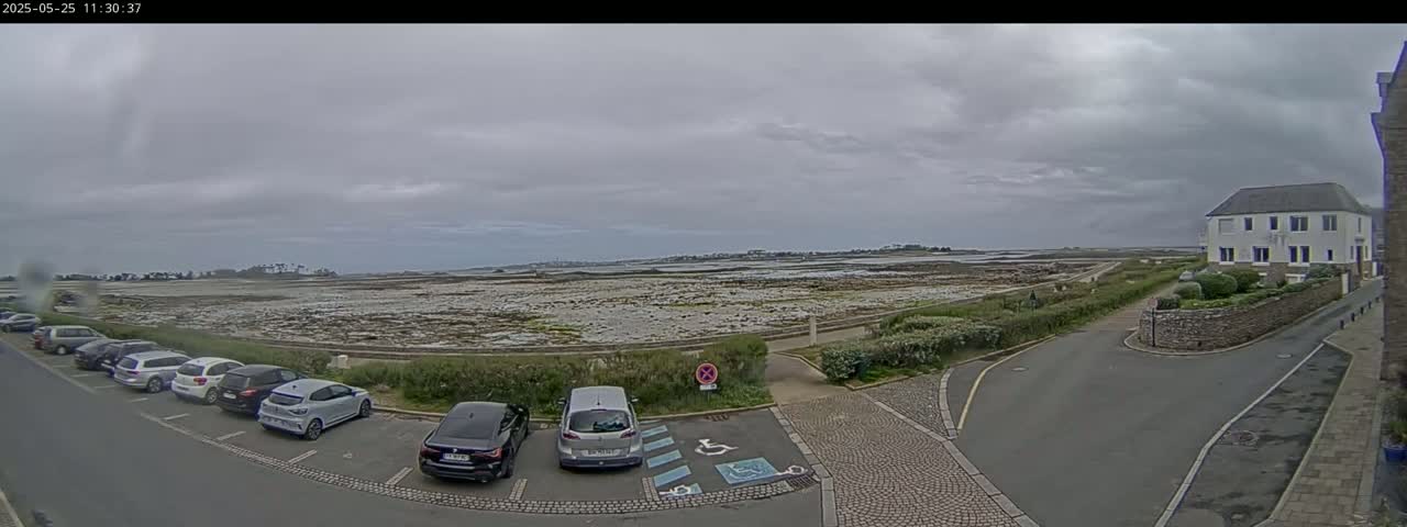 A coastal parking lot with several cars is adjacent to a low-lying expanse of rocky land under a cloudy sky.