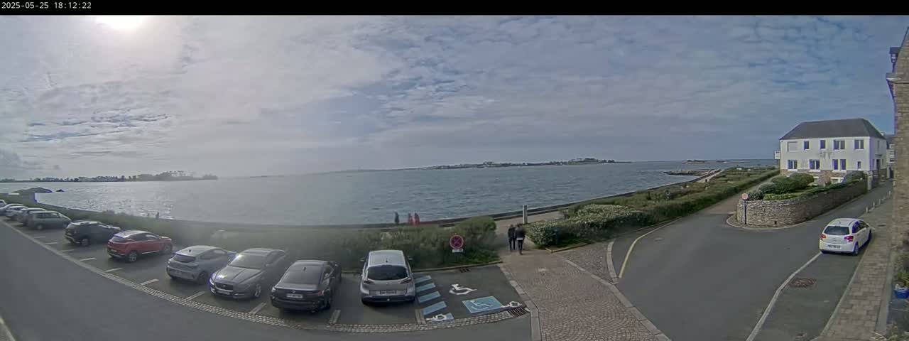 A partly cloudy day overlooks a calm body of water with several cars parked in a lot near a building.