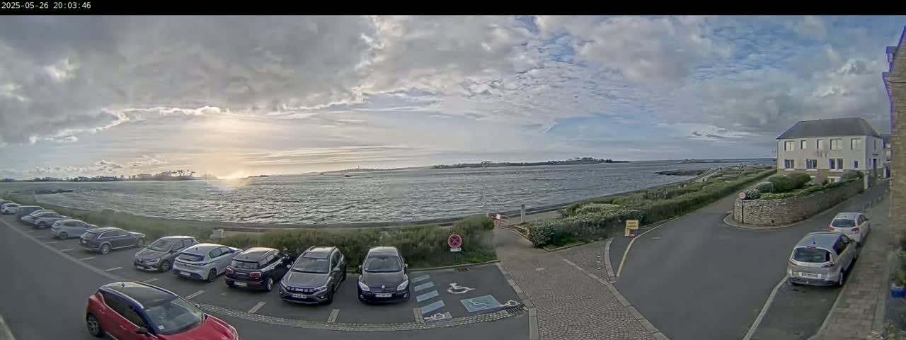 A parking lot with several cars is situated near a body of water under a partly cloudy sky, with a building visible in the background.