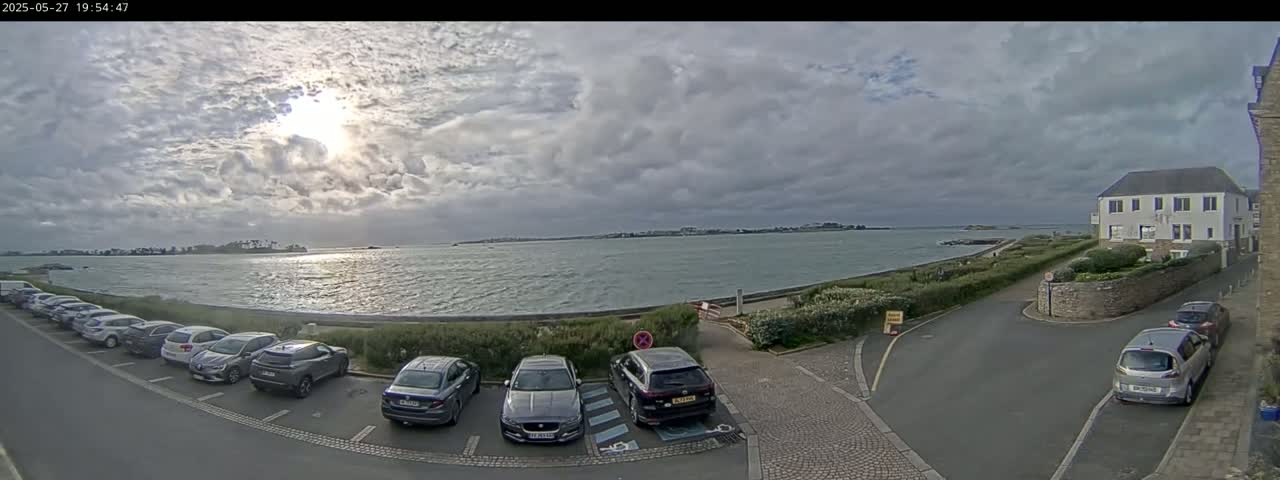 A partly cloudy day shows a body of water with a distant shoreline, several cars parked in a lot near a building, and a street leading towards the water.