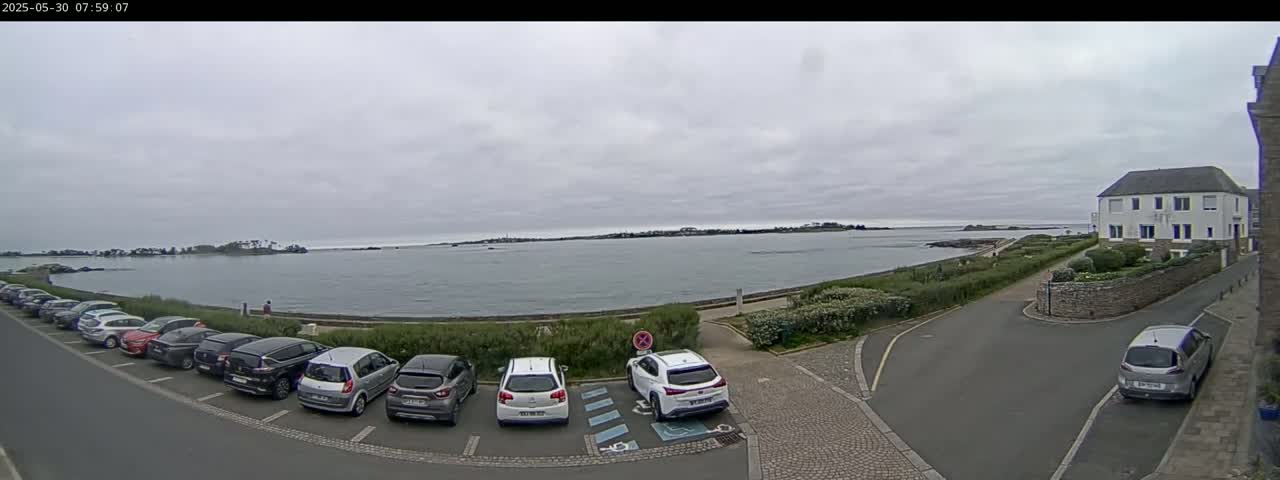 A parking lot filled with cars overlooks a calm body of water under an overcast sky.
