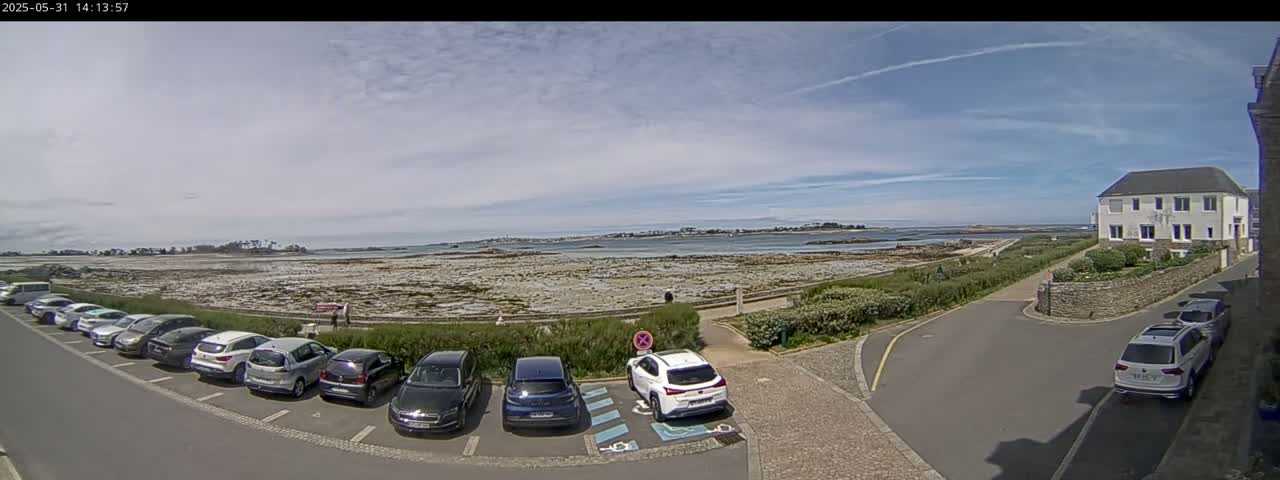 A parking lot filled with cars is situated near a coastal area under a partly cloudy sky, with a body of water and distant land visible in the background.