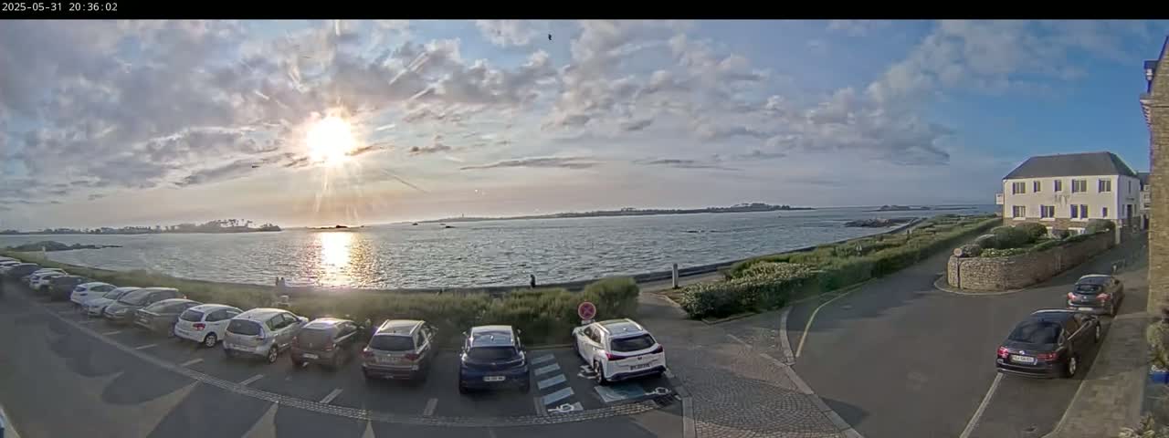 A partly sunny day shows a calm body of water with a distant shoreline, a parking lot full of cars, and a building near a stone wall, under a partly cloudy sky.