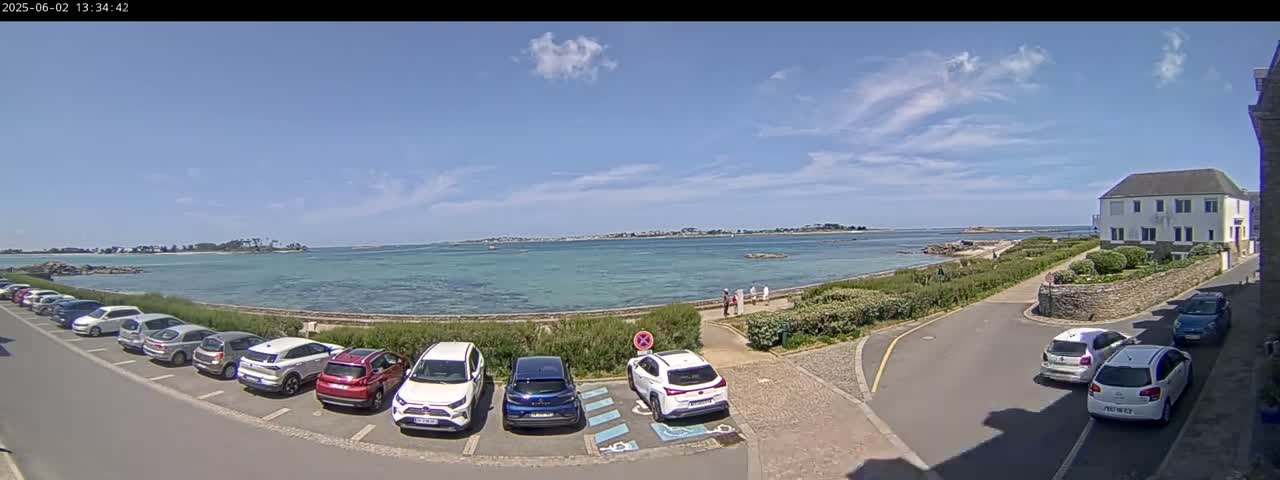 A seaside parking lot with several cars parked, overlooking a calm body of water under a mostly sunny sky.