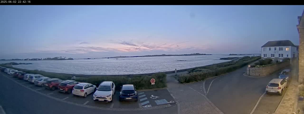 A row of cars is parked in a lot next to a calm body of water under a twilight sky, near a building and a stone wall.