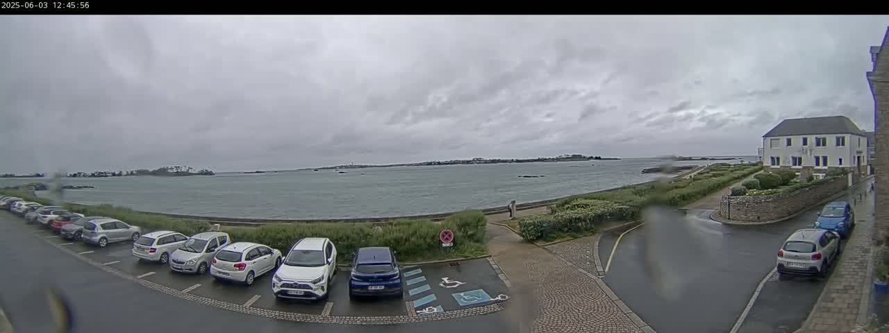 A parking lot with several cars is situated next to a body of water under an overcast sky, with a building visible in the distance.