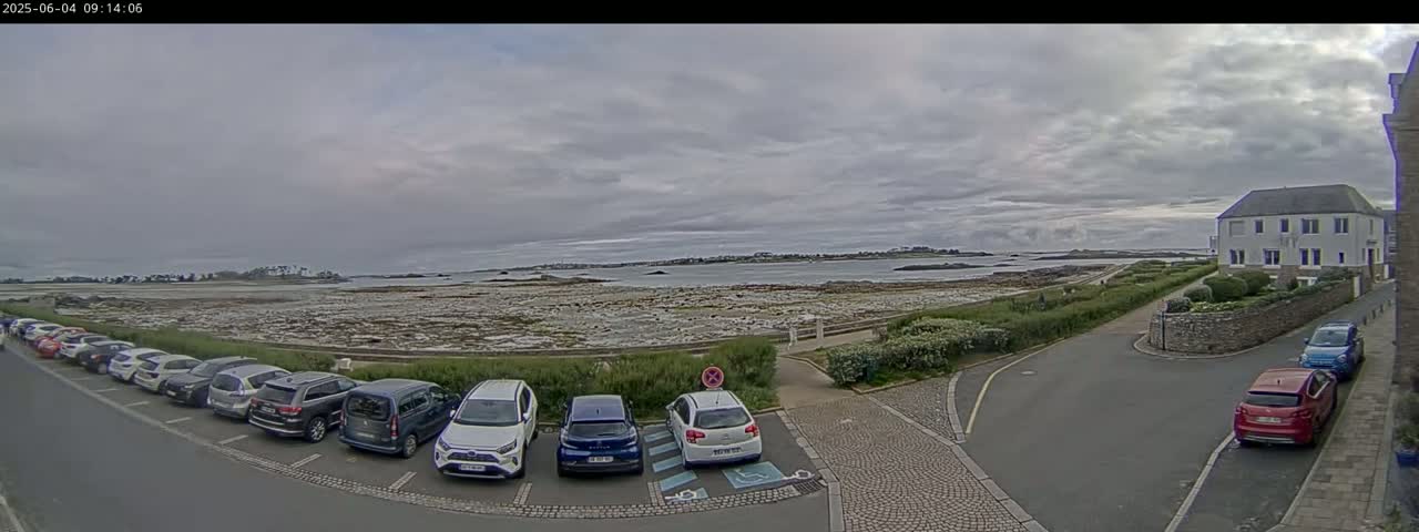 A parking lot filled with cars overlooks a rocky shoreline and calm body of water under an overcast sky.