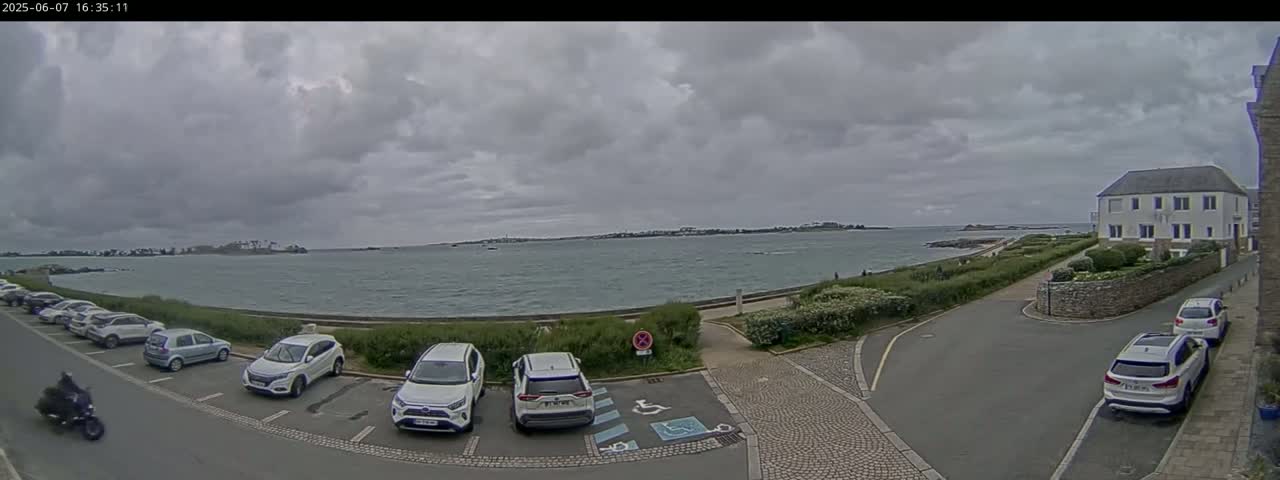 A parking lot with several cars and a motorcycle is situated next to a calm body of water under an overcast sky, with a building and greenery visible in the background.