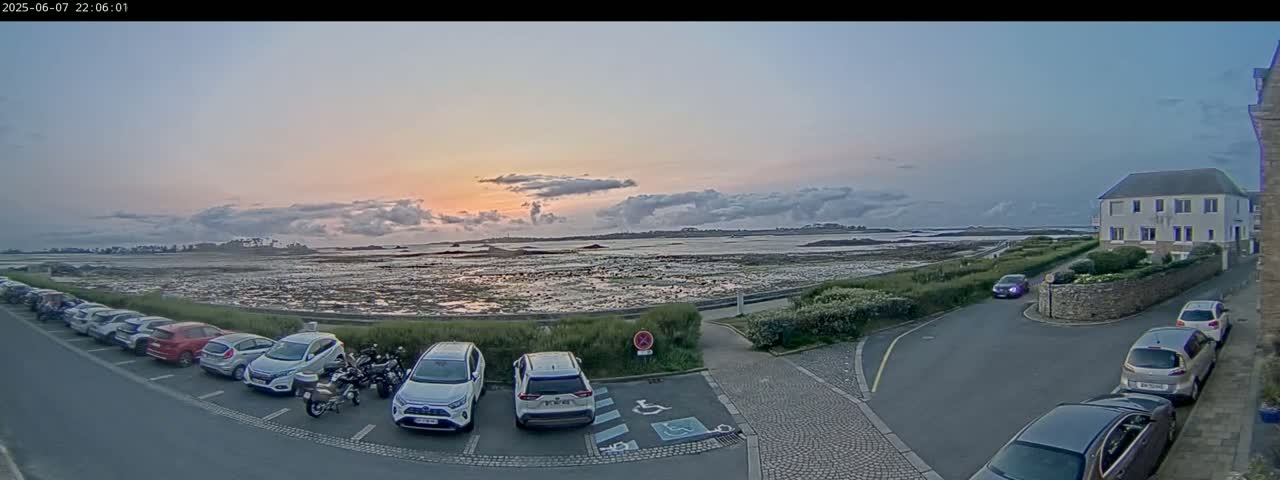 A coastal parking lot with several cars is visible at sunset, overlooking a calm body of water and low-lying land under a partly cloudy sky.
