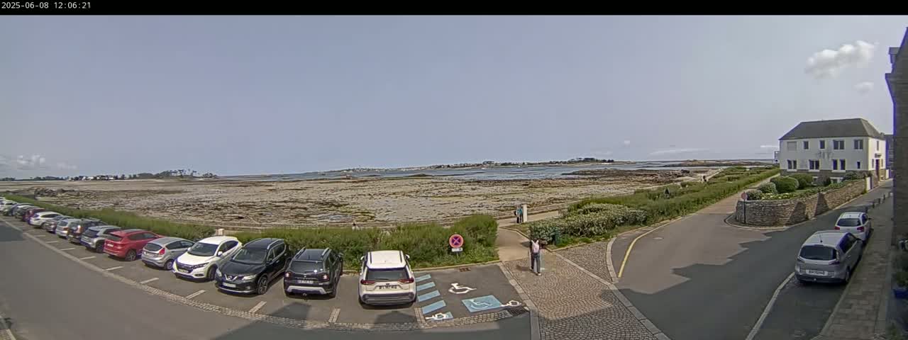 A parking lot filled with cars is situated near a shoreline with exposed rocks under a partly cloudy sky.