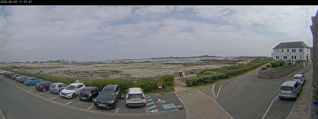 A parking lot with several cars is situated near a low-lying, rocky coastline under a mostly cloudy sky.