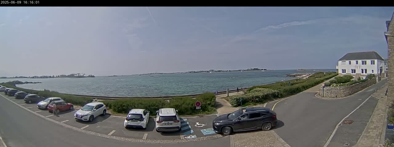 A parking lot with several cars is situated near a calm body of water on a sunny day, with a building and some greenery visible in the background.