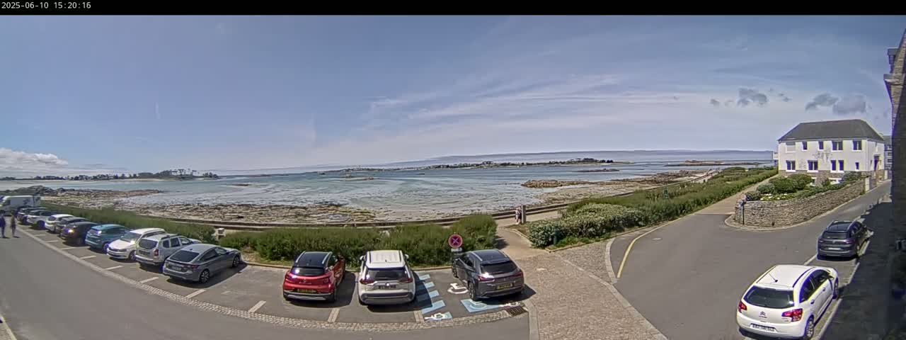 A parking lot with several cars is situated near a calm body of water under a mostly clear sky, with a shoreline and a white building visible in the distance.