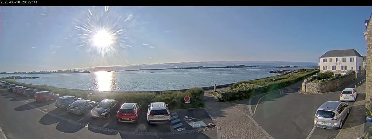 A sunny day reveals a parking lot of cars next to a calm body of water with a distant shoreline, and a building sits to the right.