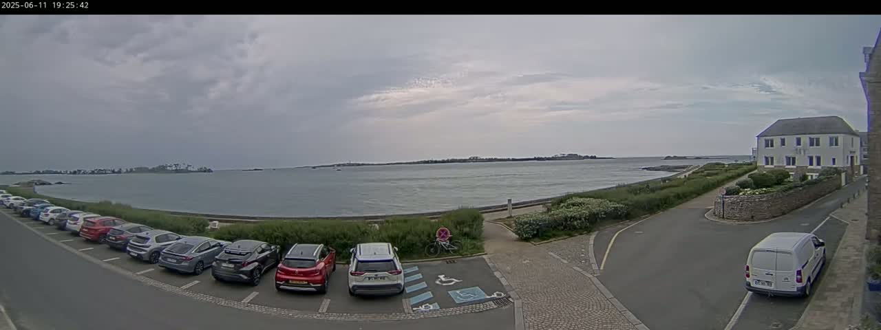 A parking lot filled with cars overlooks a calm body of water under a cloudy sky, with a building visible in the background.