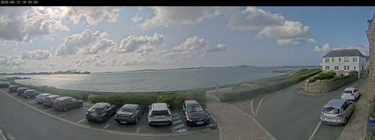 A partly sunny day shows a calm body of water with a shoreline featuring parked cars, a building, and a paved road.