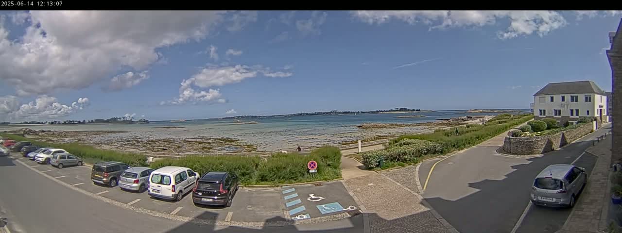 A partly cloudy sky overlooks a coastal parking lot with several cars parked near a building and a shoreline with exposed rocks and shallow water.