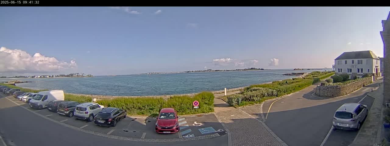 A partly sunny day shows a parking lot near a body of water with several cars parked and a building in the background.