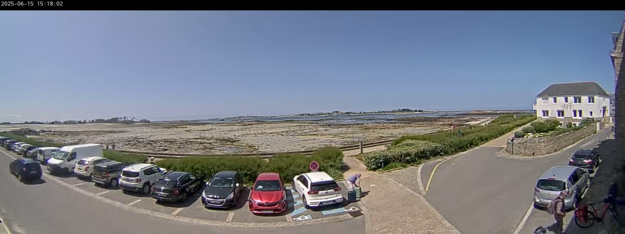 A parking lot with several cars is situated near a coastal area under a clear, sunny sky; a low stone wall separates the parking area from a flat expanse of sand and water.