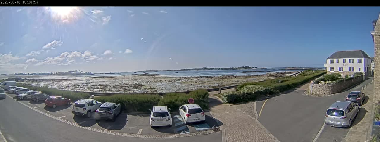 A sunny day reveals a coastal parking lot with several cars, overlooking a rocky shoreline and calm ocean water, with a building visible in the background.
