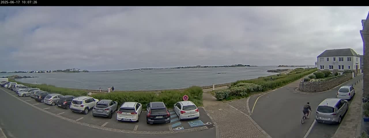 A parking lot filled with cars overlooks a calm body of water under a cloudy sky, with a person on a bicycle and a building visible in the background.