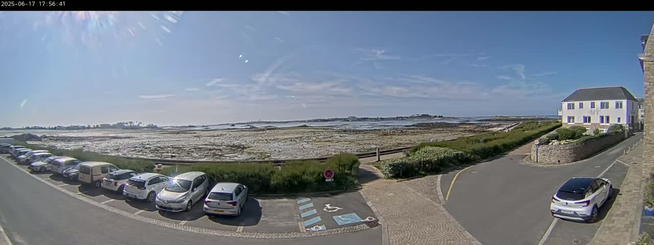 A sunny day reveals a coastal parking lot with several cars, a low stone wall separating it from a rocky, tidal shoreline, and a white building in the background.