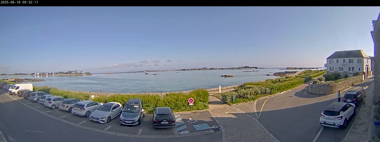 A clear, sunny day overlooks a calm body of water with several parked cars lining a street next to a low stone wall and a building.