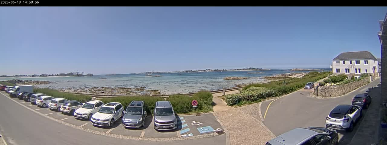 A seaside parking lot with several cars is next to a calm body of water under a clear blue sky, and a light-colored building is visible in the background.