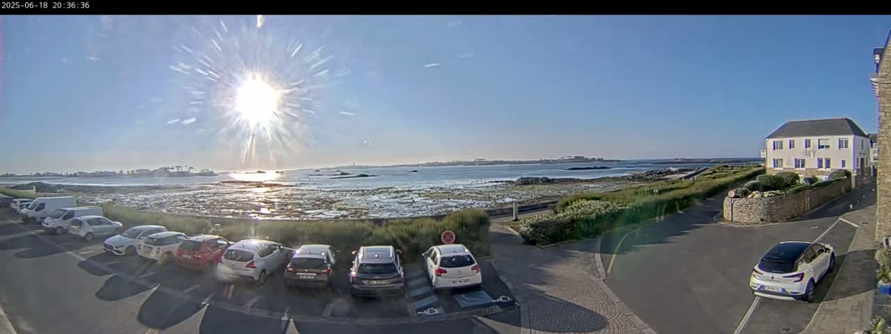 A sunny day reveals a coastal scene with a parking lot full of cars near a building overlooking the calm ocean and rocky shoreline.