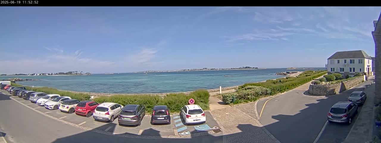 A seaside parking lot with several cars is next to a calm, blue ocean on a sunny day, with a distant shoreline and a white building visible.