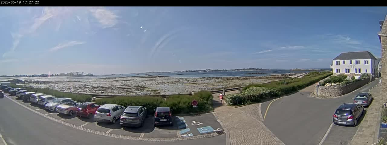 A sunny day reveals a coastal scene with a parking lot full of cars near a building overlooking a calm body of water and exposed rocky shoreline.