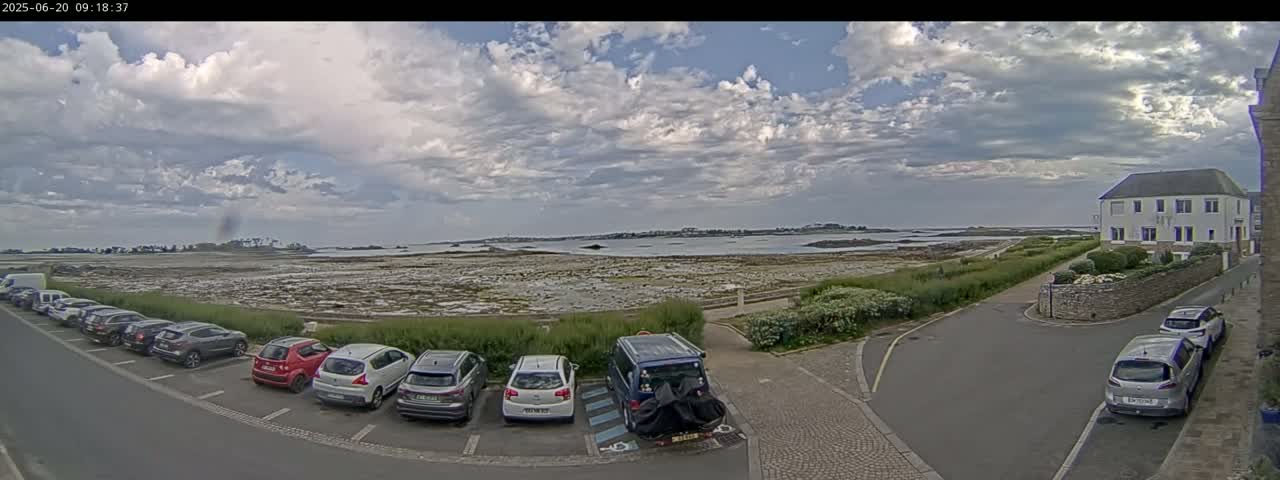 A parking lot with several cars is situated near a coastal area with a calm body of water under a partly cloudy sky.