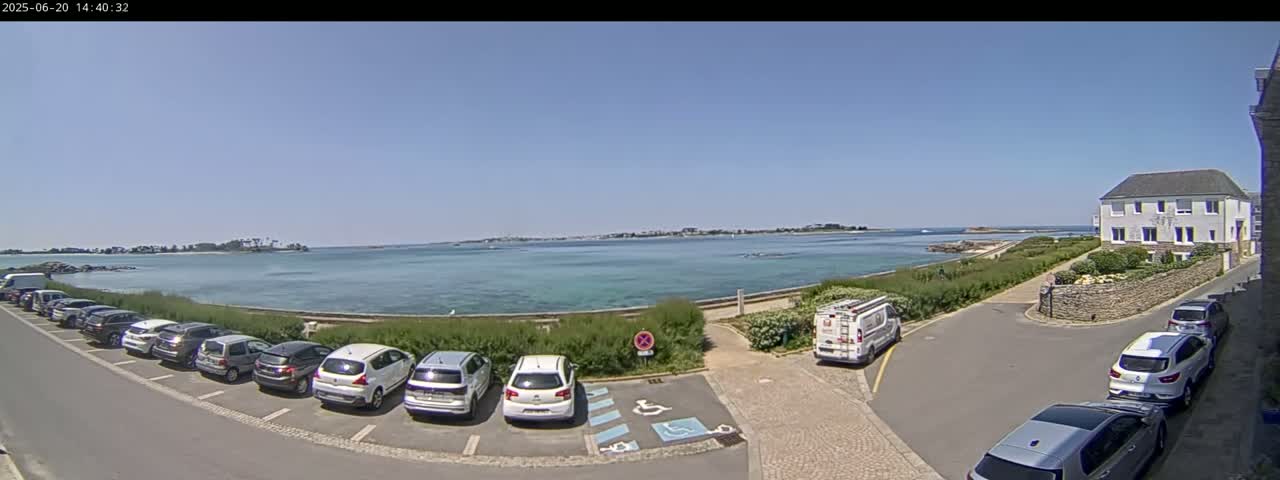 A parking lot with several cars is situated next to a calm, light-blue body of water under a clear, sunny sky, with a building and distant land visible in the background.