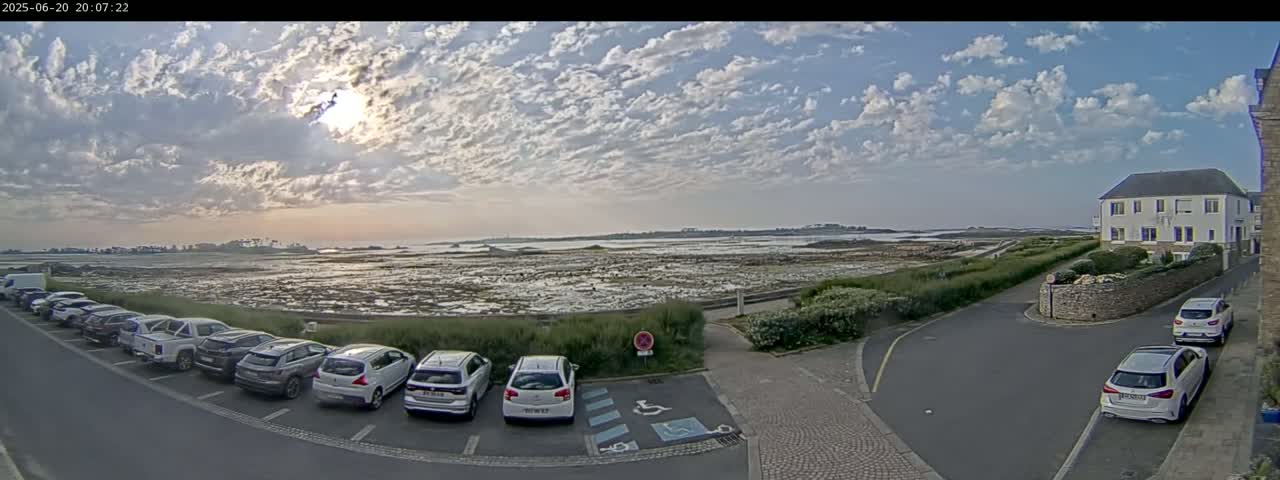 A partly cloudy sunset over a tidal flat and ocean, with a parking lot full of cars and a building visible in the foreground.