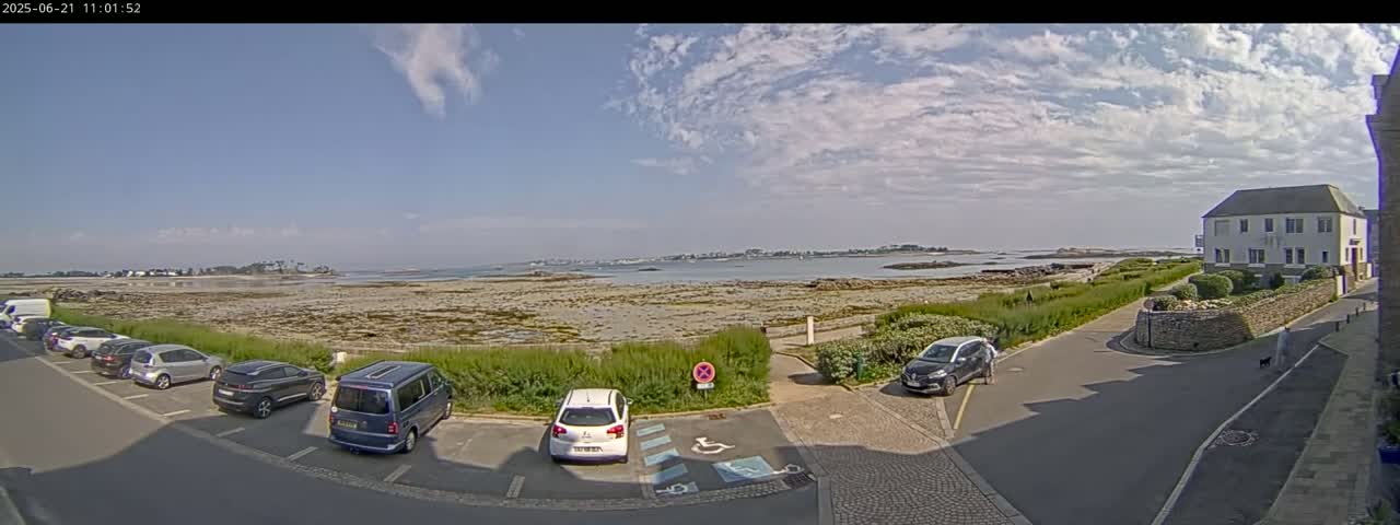 A partly cloudy sky overlooks a coastal scene with several cars parked in a lot near a building and a wide expanse of beach leading to calm water and distant land.