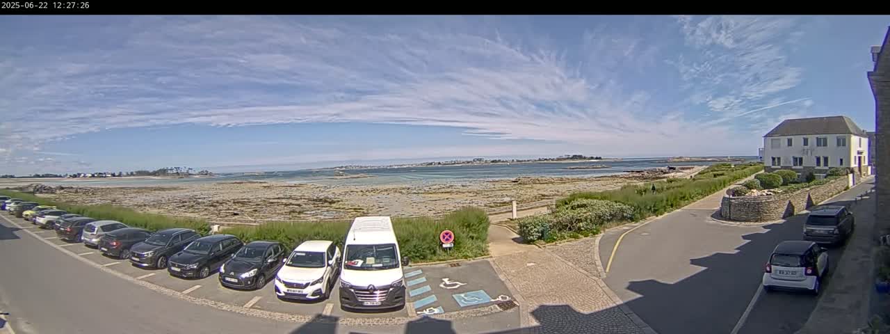 A partly sunny day reveals a parking lot filled with cars next to a calm body of water and a shoreline, with a building visible in the background.