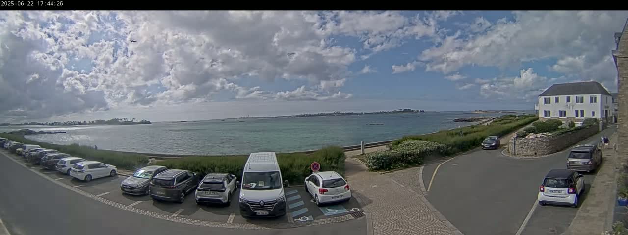 A partly cloudy sky overlooks a calm body of water with several cars parked in a lot near a building and a road.