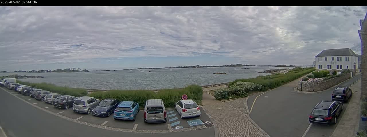 A partly cloudy day reveals a calm body of water with a distant shoreline, overlooked by a parking lot filled with cars and a residential building.
