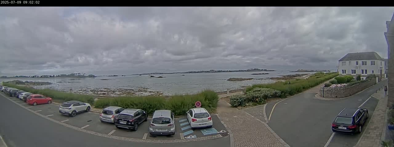 A parking lot with several cars is next to a coastal road overlooking a calm body of water under a cloudy sky.